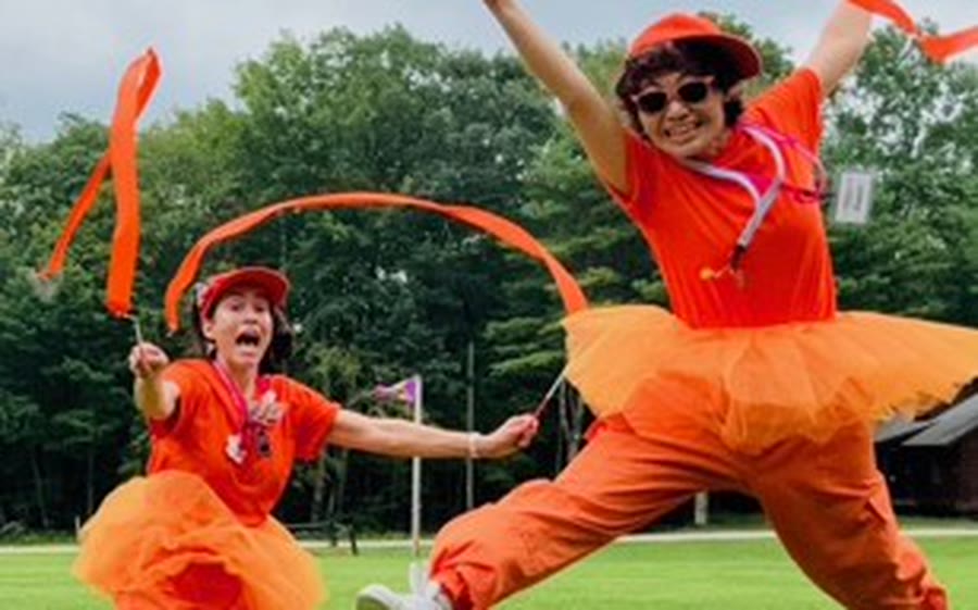 Two Red Nose Docs in orange leap across a camp lawn trailing ribbon streamers.