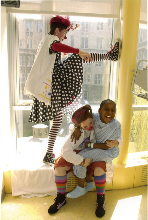 Two Red Nose Docs play with a smiling young patient on a sunlit hospital windowsill, one balanced against the window frame.