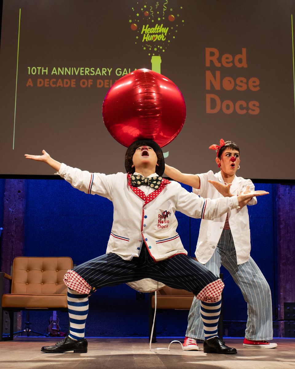 Two Red Nose Docs perform on stage in white coats and striped trousers, one balancing a large red balloon on their head with both arms flung wide, in front of a screen reading A Decade of Delight &mdash; Red Nose Docs.