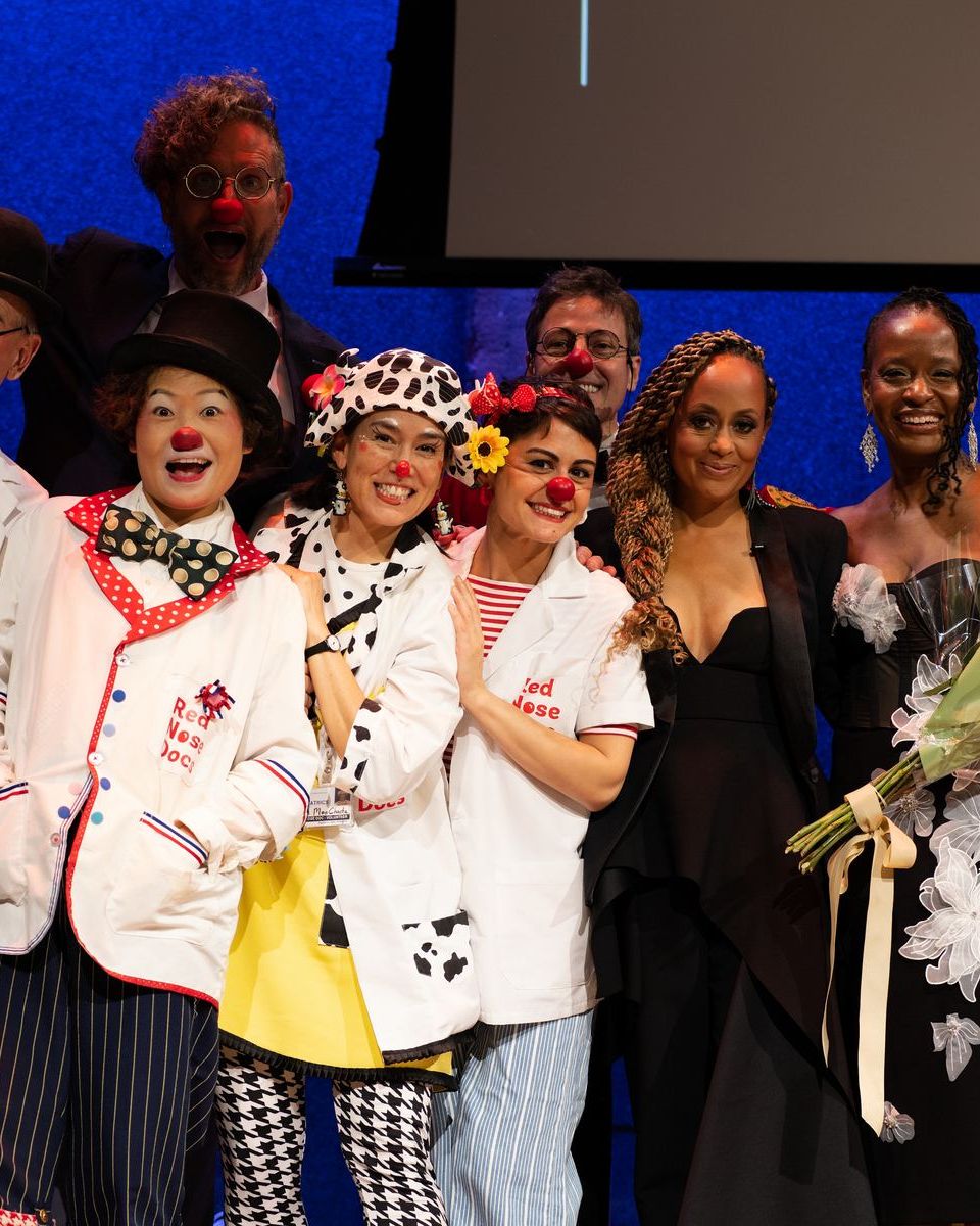 The Red Nose Docs crowd together on stage at the end of the evening, in lab coats and red noses, beside host Essence Atkins holding a bouquet.