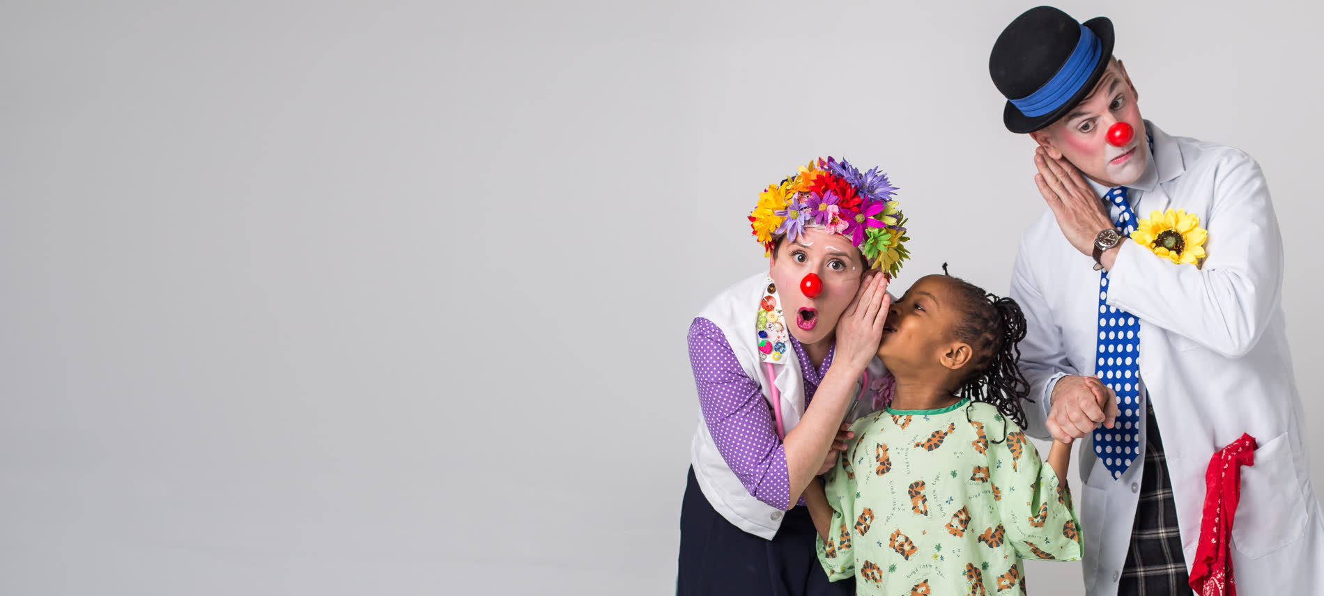 A child in a hospital gown whispers to a Red Nose Doc while a second Red Nose Doc leans in to listen.
