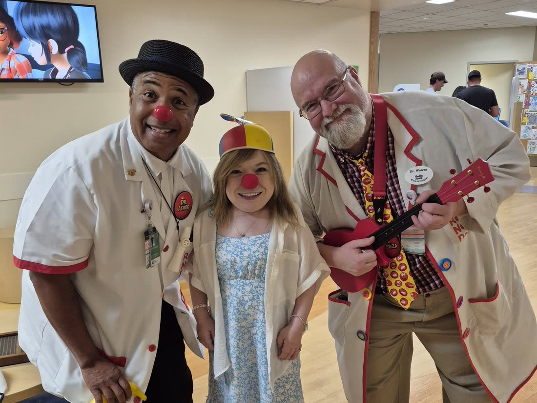 Two Red Nose Docs, one holding a ukulele, with a young patient wearing a red nose.