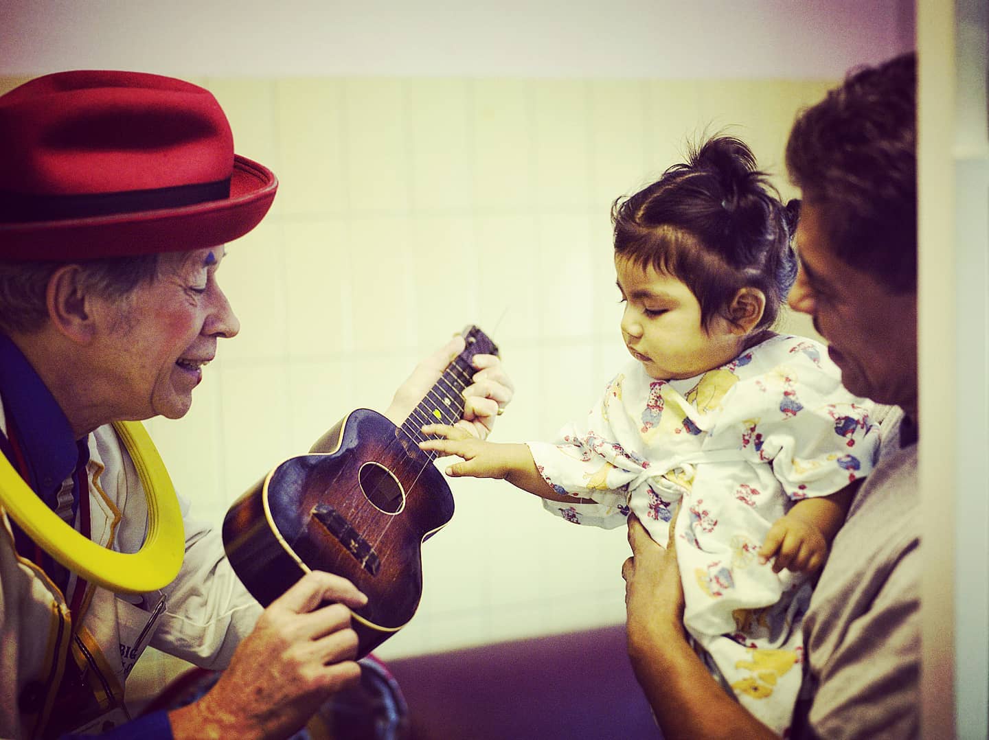 A Red Nose Doc plays a ukulele at the bedside as a toddler in a hospital gown reaches out to touch the strings.