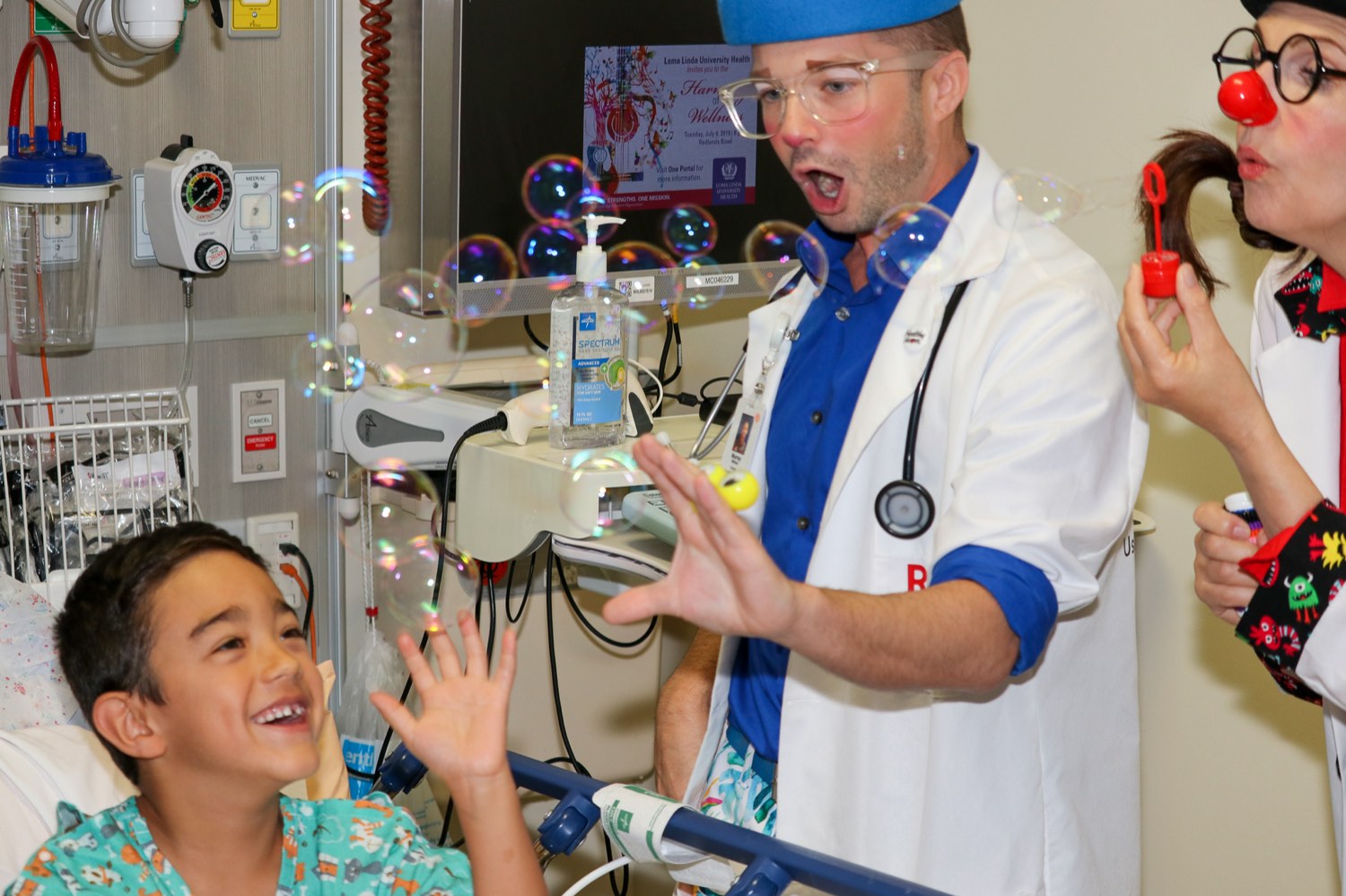 A Red Nose Doc fills a hospital room with bubbles as a young patient laughs.