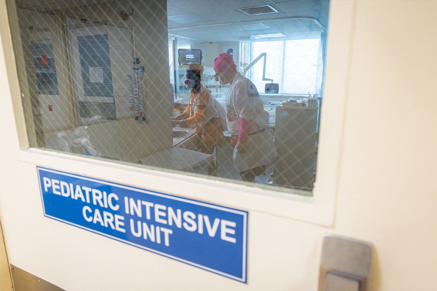 Two gowned Red Nose Docs at work inside a pediatric intensive care unit, seen through the unit window.